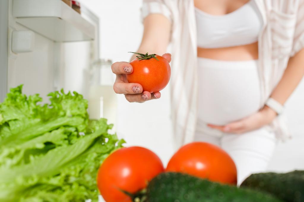 Foco seletivo da mulher grávida segurando tomate perto de legumes frescos no refrigerador aberto isolado no branco
 - Foto, Imagem