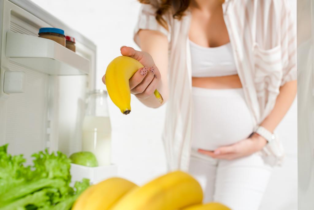 Selective focus of pregnant woman holding banana in open fridge on white background - Photo, Image