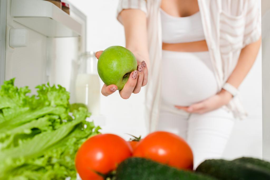 Viljelty näkymä raskaana oleva nainen tilalla omena lähellä tuoreita vihanneksia avoimessa jääkaapissa valkoisella pohjalla
 - Valokuva, kuva