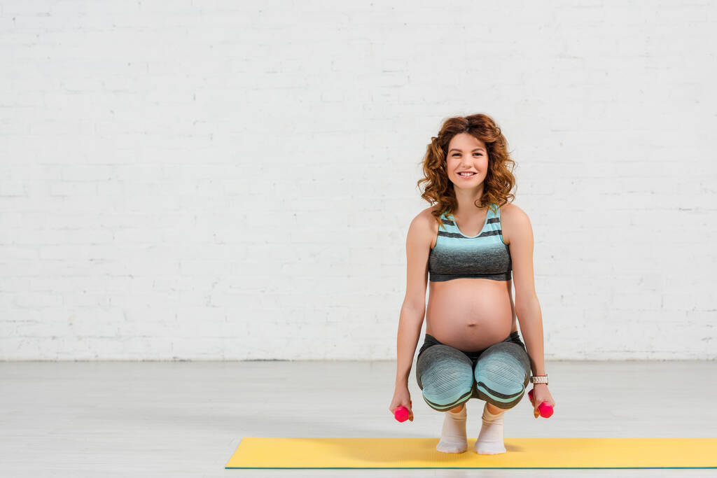 Έγκυος γυναίκα σε αθλητικά ρούχα χαμογελώντας στην κάμερα και την κατάρτιση με αλτήρες στο στρώμα γυμναστικής στο σπίτι - Φωτογραφία, εικόνα