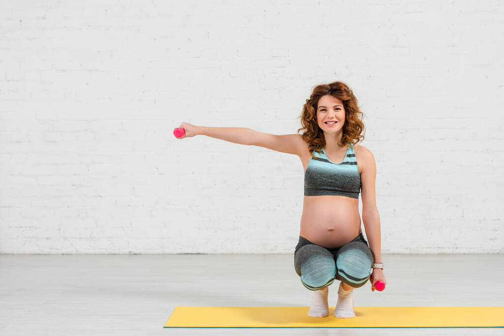 Schwangere lächelt in die Kamera und trainiert zu Hause mit Kurzhanteln auf Fitnessmatte - Foto, Bild