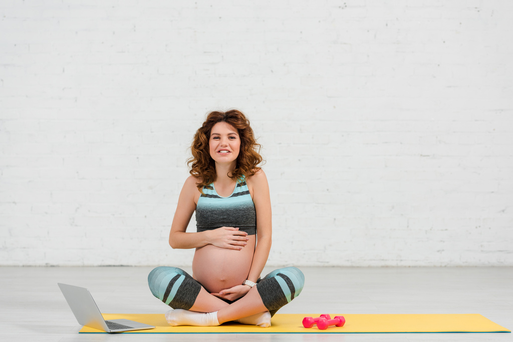 Schwangere lächelt in die Kamera und berührt Bauch in der Nähe von Laptop und Hanteln auf Fitnessmatte - Foto, Bild