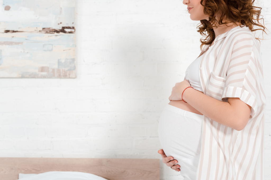Обрезанный вид беременной женщины, трогающей животик в спальне
 - Фото, изображение