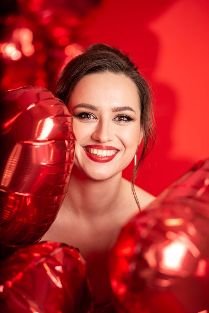 Schöne junge Frau in rotem Abendkleid posiert vor rotem Hintergrund mit großen herzförmigen Luftballons.  - Foto, Bild