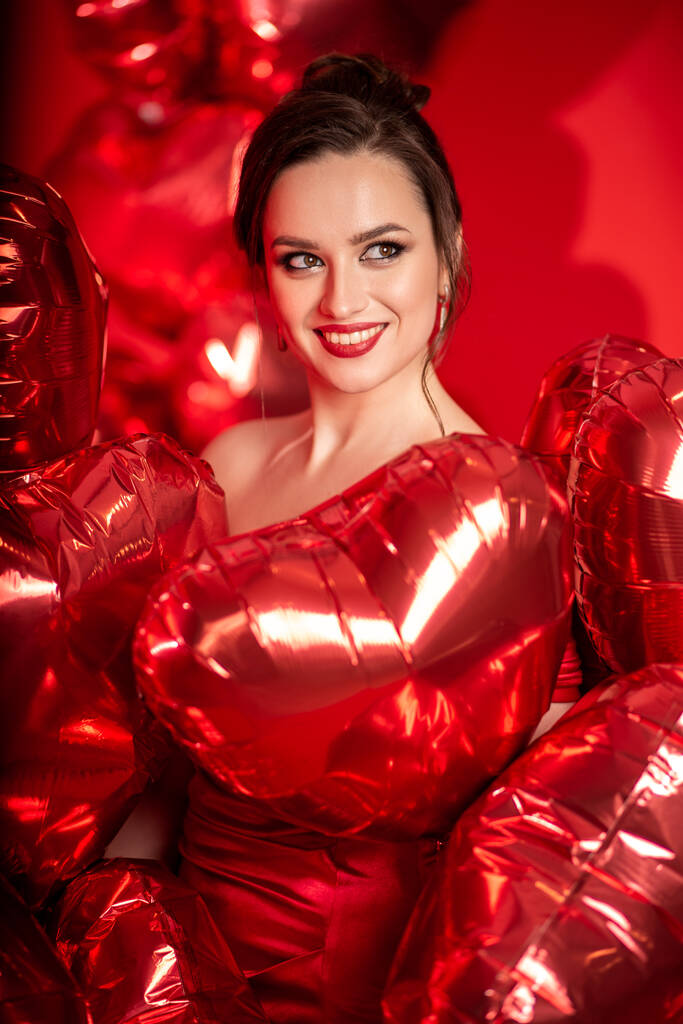 Красива молода жінка в червоній вечірній сукні позує на червоному тлі з кульками великої форми серця
.  - Фото, зображення