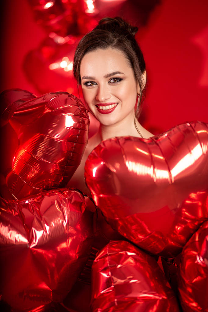 Belle jeune femme en robe de soirée rouge posant sur fond rouge avec de grands ballons en forme de coeur
.  - Photo, image
