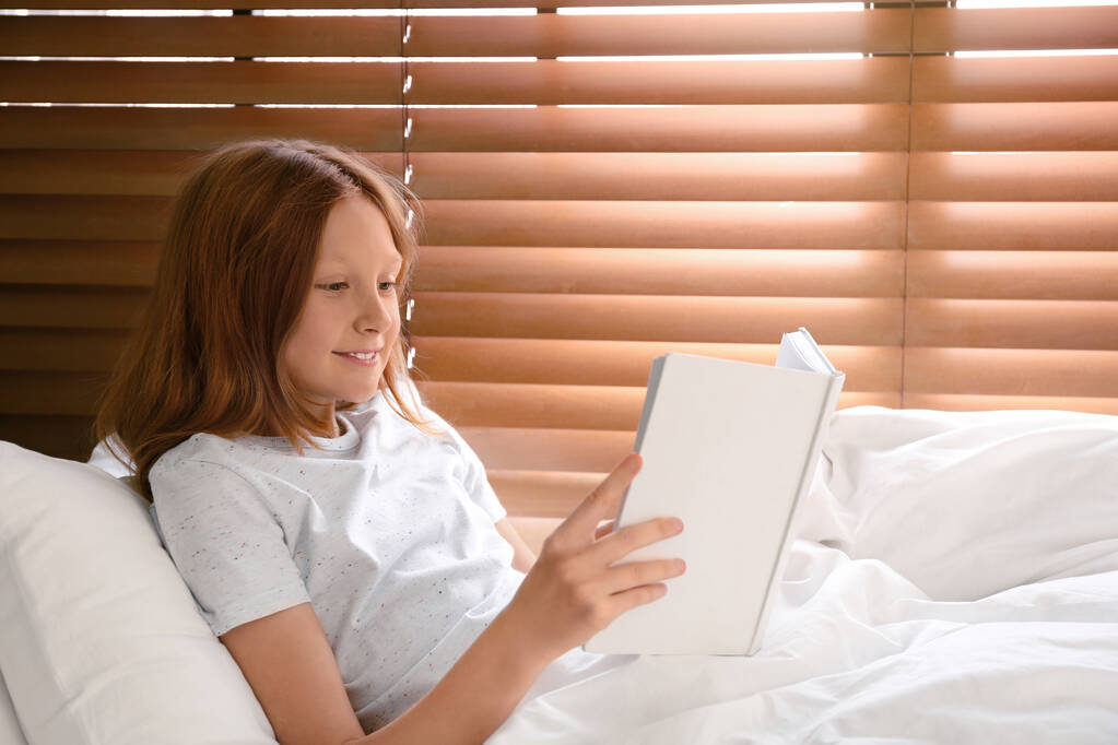 Милая девочка-подросток читает книгу в постели у окна
 - Фото, изображение