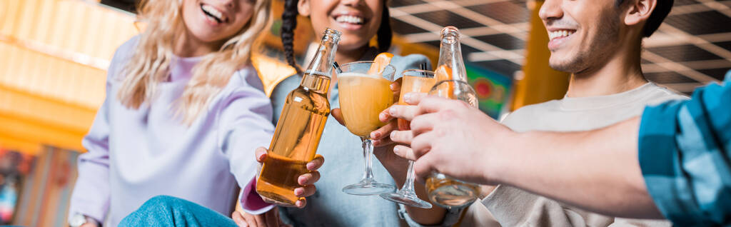abgeschnittener Blick auf fröhliche multikulturelle Freunde, die mit Bierflaschen und Cocktailgläsern klappern, während sie im Bowlingclub sitzen, Panoramaaufnahme - Foto, Bild