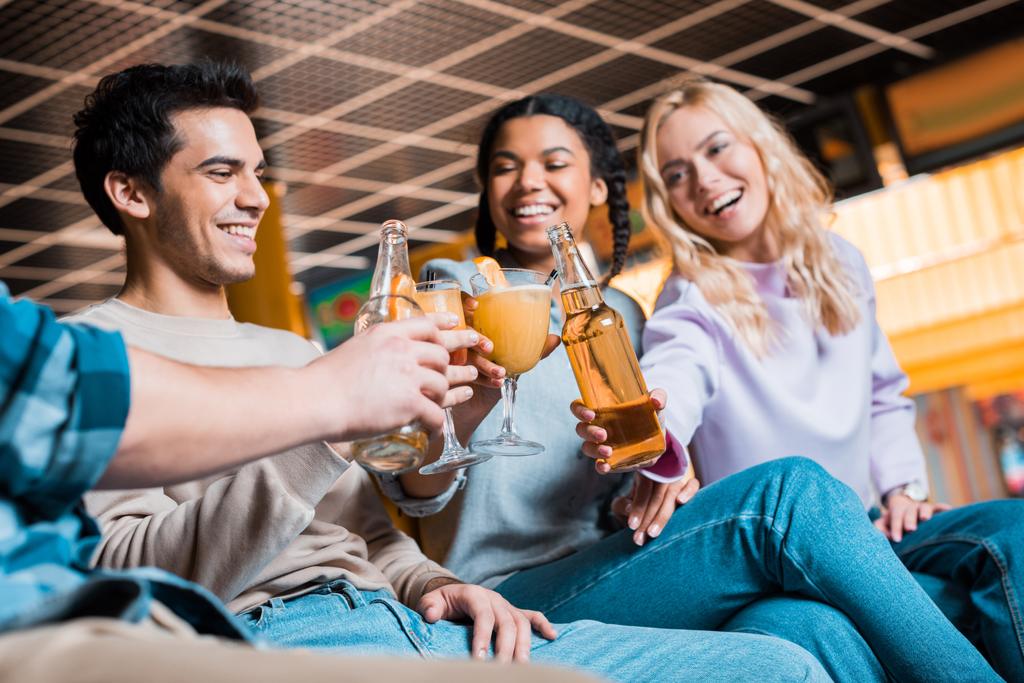 cheerful multicultural friends clinking with bottles of beer and cocktail glasses while sitting in bowling club  - Photo, Image