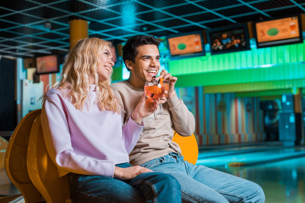vrolijk paar kijken weg en glimlachen tijdens het drinken van cocktails in bowling club - Foto, afbeelding