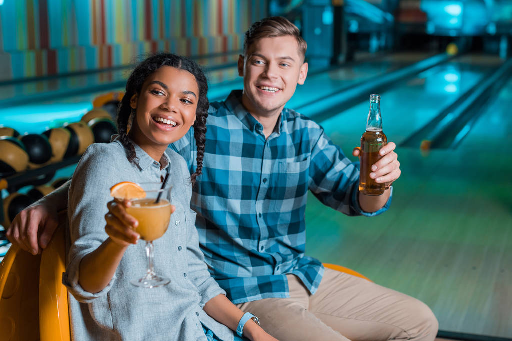 glückliches gemischtrassiges Paar schaut weg, während es mit Cocktail und Bier im Bowlingclub sitzt - Foto, Bild