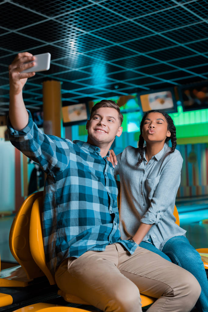 cheerful interracial couple taking selfie on smartphone while sitting in bowling club - Photo, Image