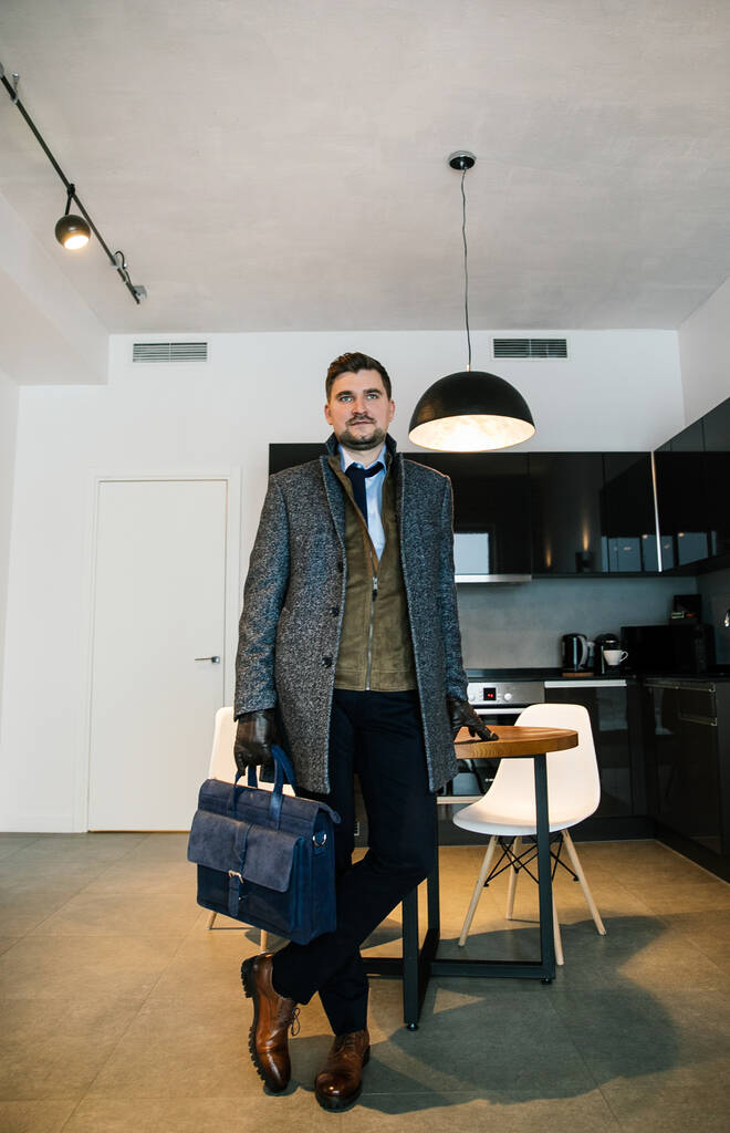 modisch gekleideter Geschäftsmann mit Bart - schicker Anzug, braune Schuhe, Krawatte, Handschuhe, ein Mann posiert in einer teuren Wohnung - Foto, Bild