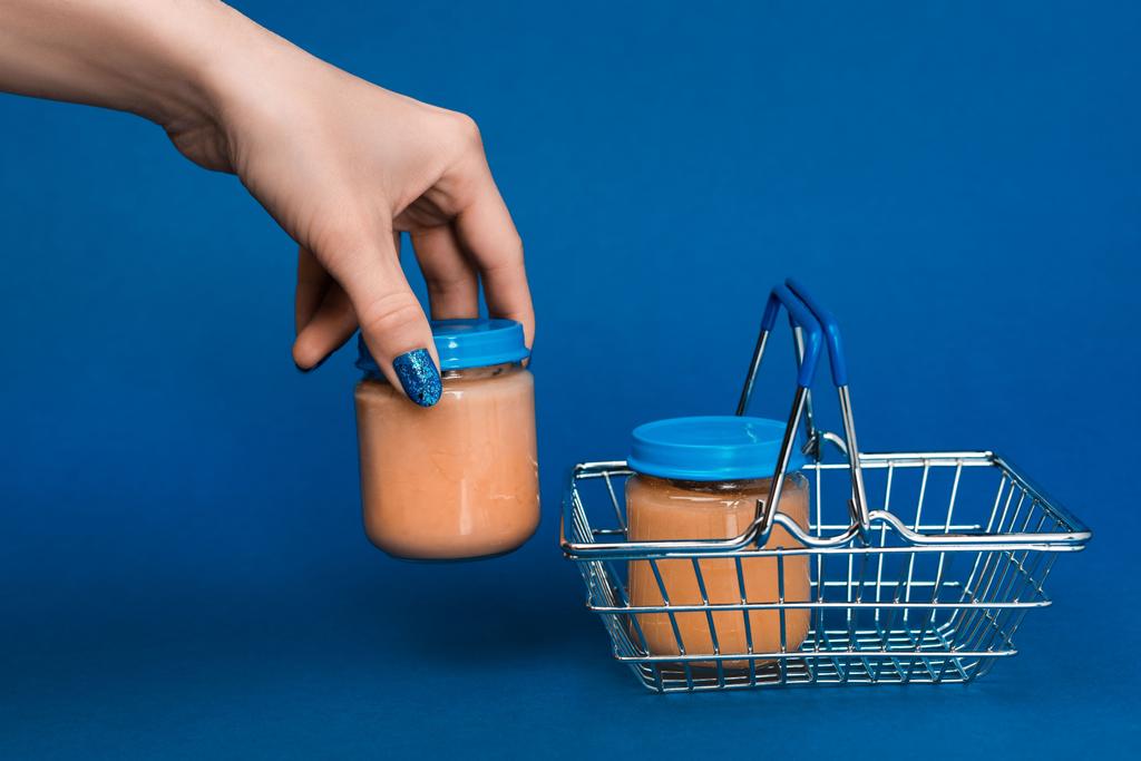 vista cortada da mulher colocando no frasco cesta de compras com comida de bebê no fundo azul
  - Foto, Imagem