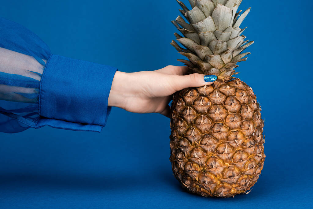 bijgesneden uitzicht van vrouw met smakelijke en hele ananas op blauwe achtergrond  - Foto, afbeelding