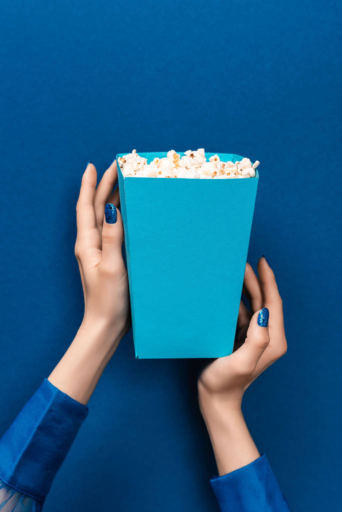 обрізаний вид жінки, що тримає коробку з попкорном на синьому фоні
  - Фото, зображення