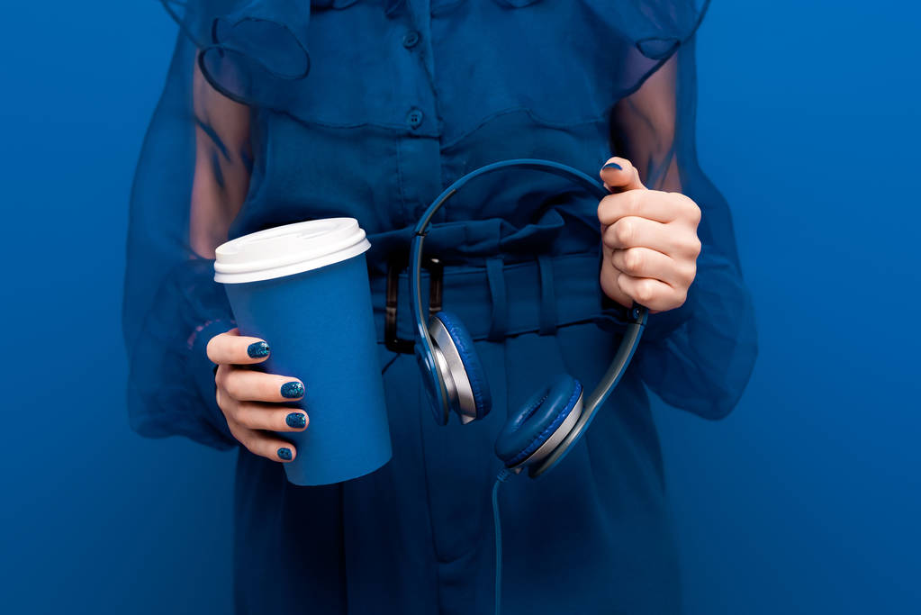 oříznutý pohled na ženu držící papírový pohár a sluchátka na modrém pozadí  - Fotografie, Obrázek