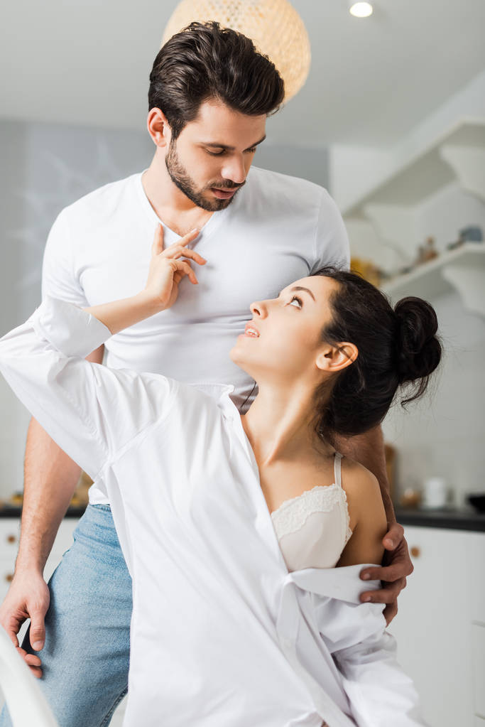 Sensual woman in shirt and bra touching chest of handsome boyfriend in kitchen - Photo, Image
