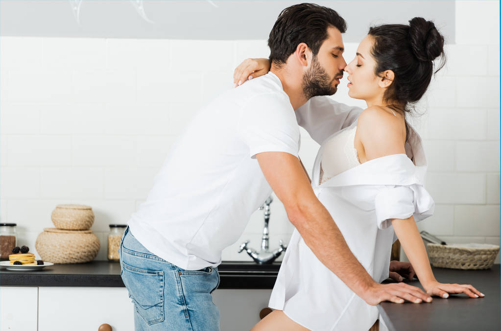 Вид сбоку мужчины, целующего чувственную женщину в бюстгальтере и рубашке на кухне
  - Фото, изображение