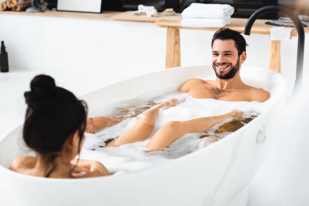 Selective focus of handsome man smiling at girlfriend in bathtub with foam - 写真・画像