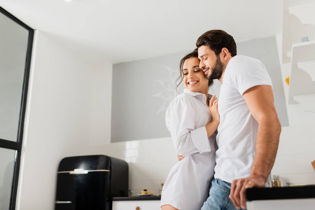 Side view of handsome man embracing smiling girlfriend in shirt near kitchen worktop - Photo, Image