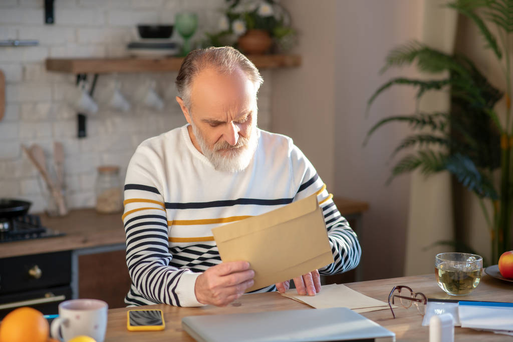 Homem barbudo de cabelos grisalhos sentado à mesa com um jornalista nas mãos
 - Foto, Imagem