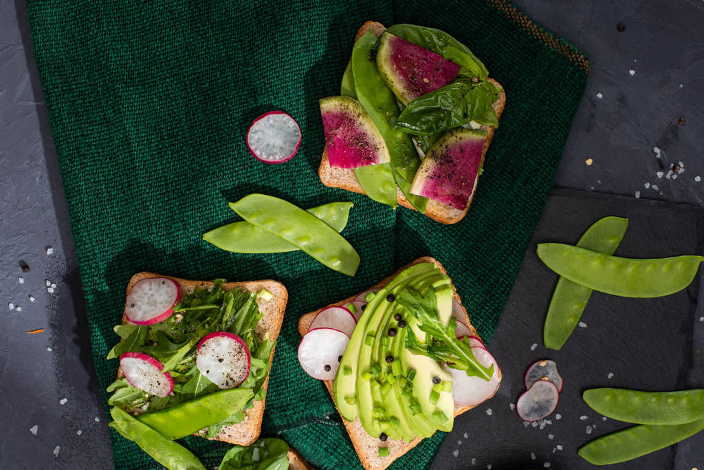 vrchní pohled na čerstvé sendviče s ředkvičkami, hráškem a avokádem na zelené látce - Fotografie, Obrázek