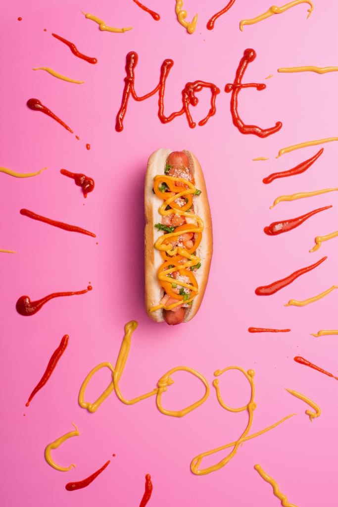 сверху вид нездорового хот-дога на розовом с словом хот-дог написан кетчупом и горчицей
 - Фото, изображение
