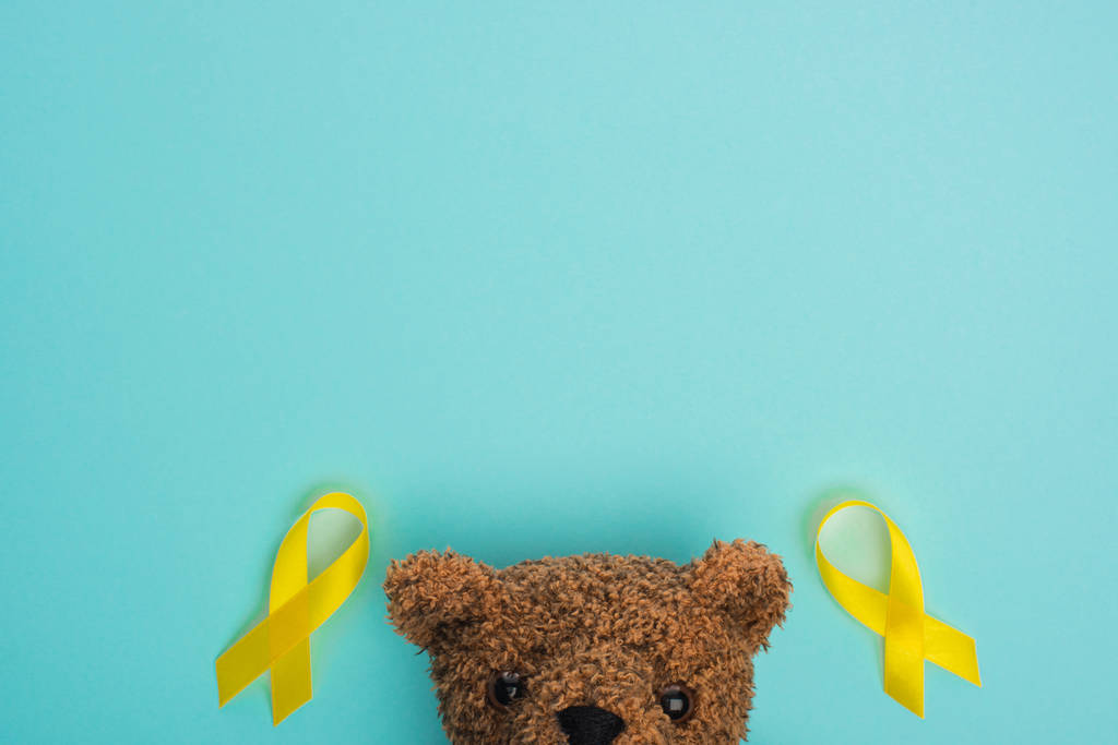 Vista superior de cintas de conciencia amarillas y oso de peluche marrón sobre fondo azul, concepto del día internacional del cáncer infantil
 - Foto, imagen