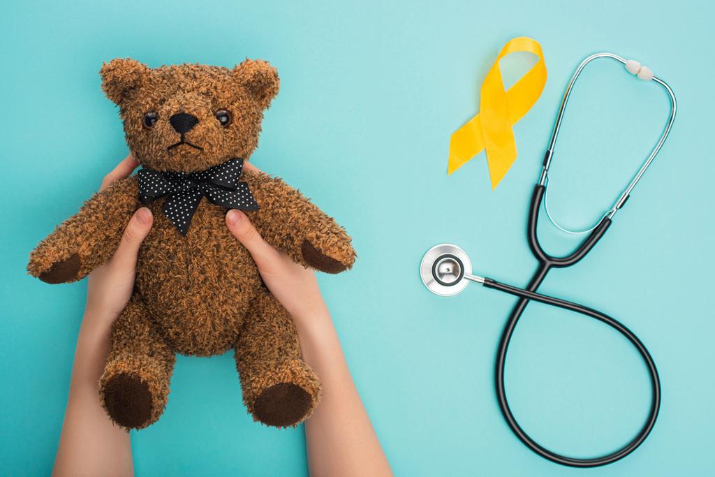 Vista recortada de la mujer sosteniendo oso de peluche cerca de cinta de conciencia amarilla y estetoscopio sobre fondo azul, concepto de día del cáncer infantil internacional
 - Foto, Imagen