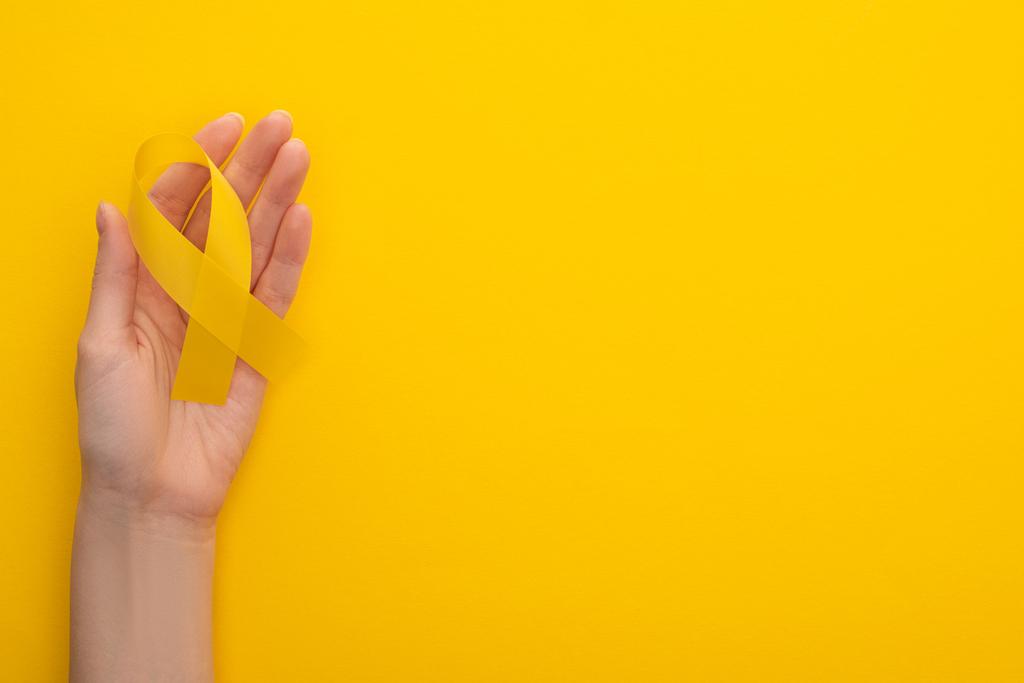 カラフルな背景に黄色のリボンを持つ女性の手の部分的なビュー,国際小児がんの日の概念  - 写真・画像