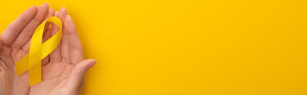 Vágott kilátás a női kéz sárga szalaggal színes háttér, panoráma lövés, nemzetközi gyermekkori rák nap koncepció - Fotó, kép