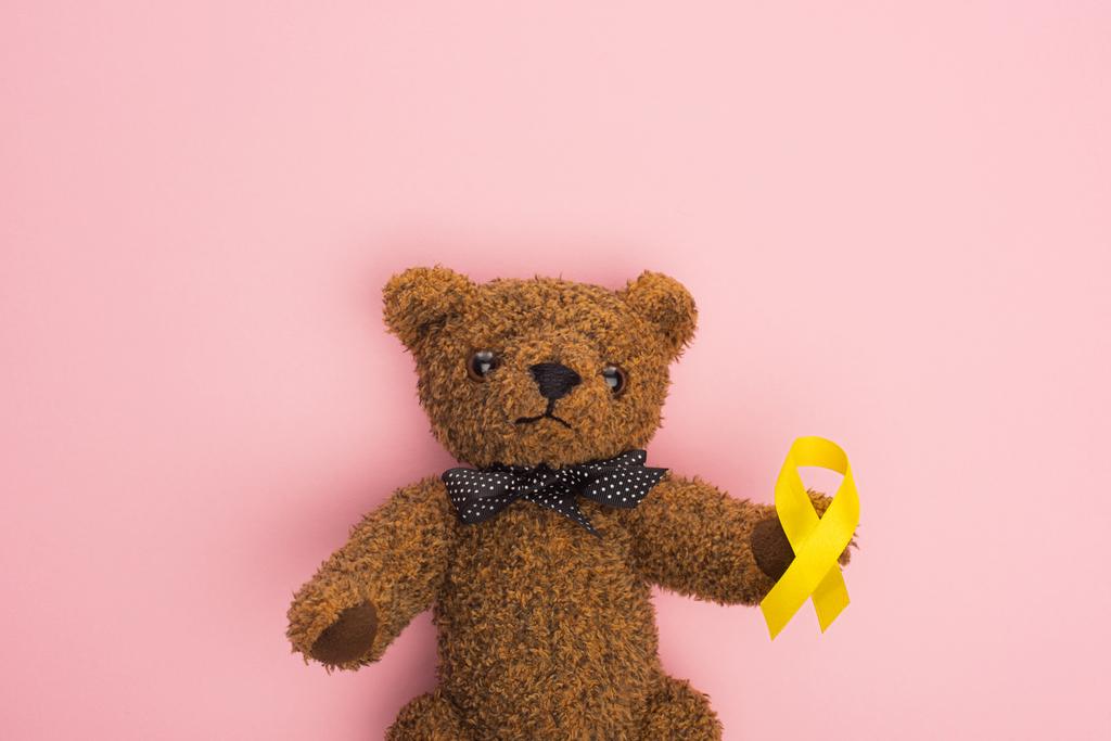 Top view of yellow ribbon on brown teddy bear with bow on pink, international childhood cancer day concept - Photo, Image