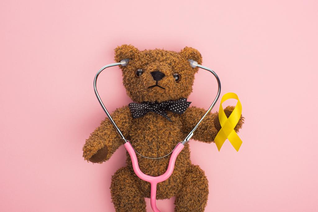 Top view of yellow ribbon and stethoscope on brown teddy bear on pink background, international childhood cancer day concept - Photo, Image