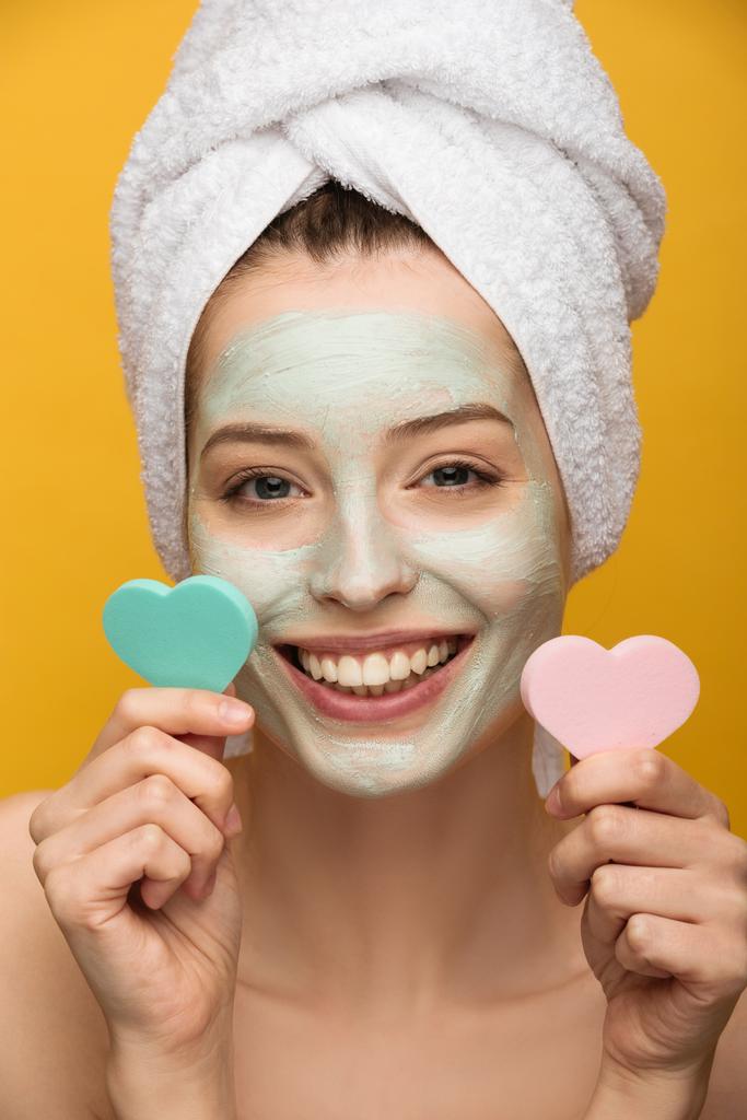 alegre chica con nutritiva máscara en la cara celebración de corazón en forma de esponjas cosméticas aisladas en amarillo
 - Foto, imagen