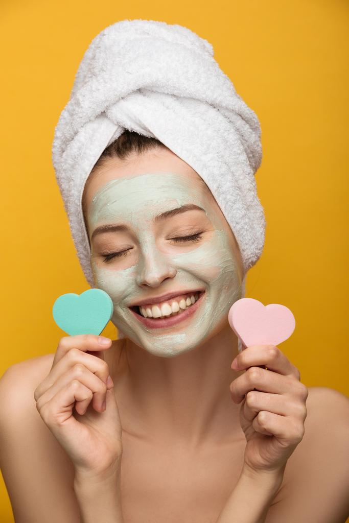 cheerful girl with nourishing facial mask holding heart-shaped cosmetic sponges with closed eyes isolated on yellow - Photo, Image