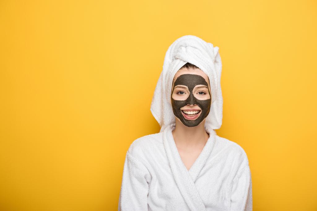 smiling girl with facial clay mask and towel on head looking at camera on yellow background - Photo, Image