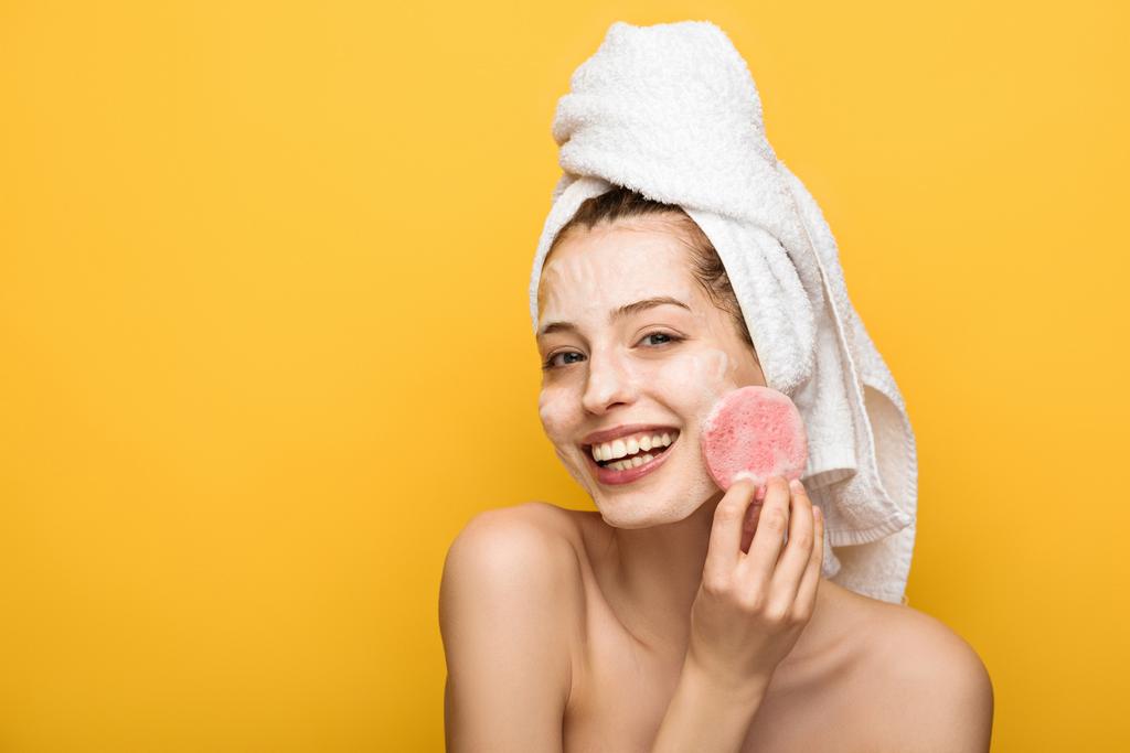 happy girl with moisturizing facial mask looking at cosmetic sponge isolated on yellow - Photo, Image