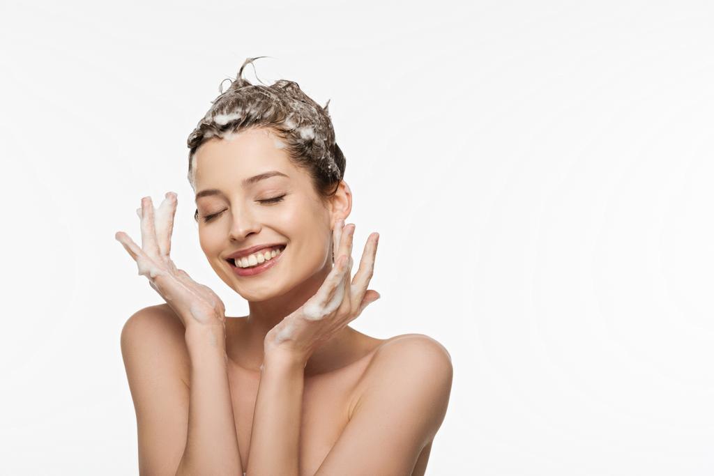 usmívající se dívka s mýdlovými vlasy a zavřenýma očima držící ruce blízko obličeje izolované na bílém - Fotografie, Obrázek