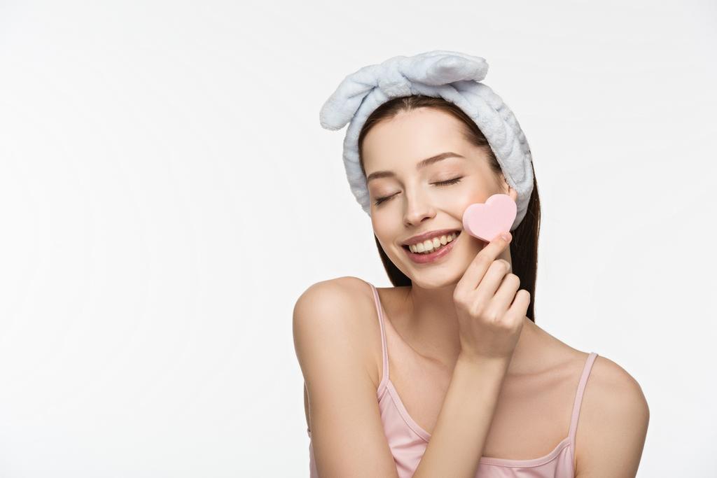 happy girl with closed eyes holding heart-shaped cosmetic sponge near face isolated on white - Photo, Image