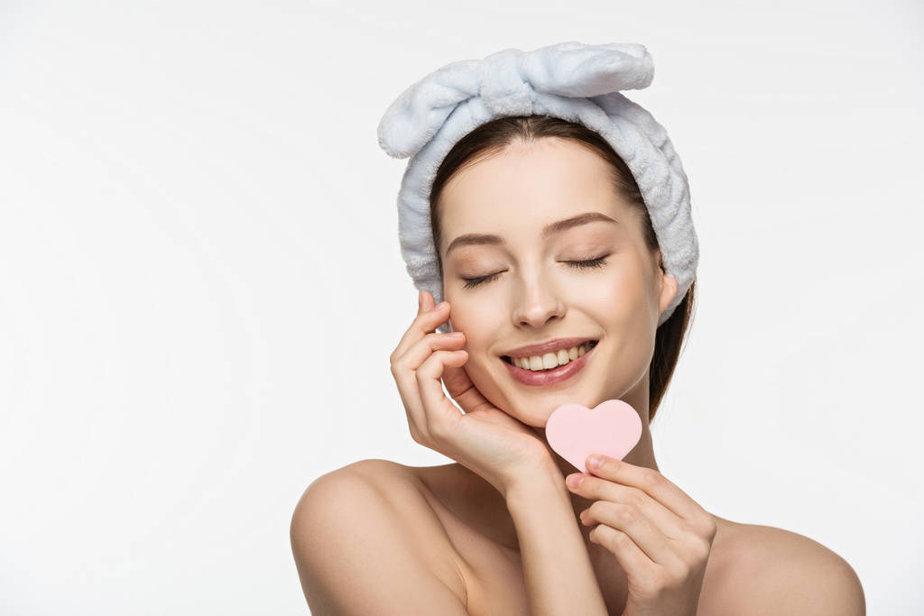 happy girl with closed eyes holding heart-shaped cosmetic sponge while touching face isolated on white - Photo, Image