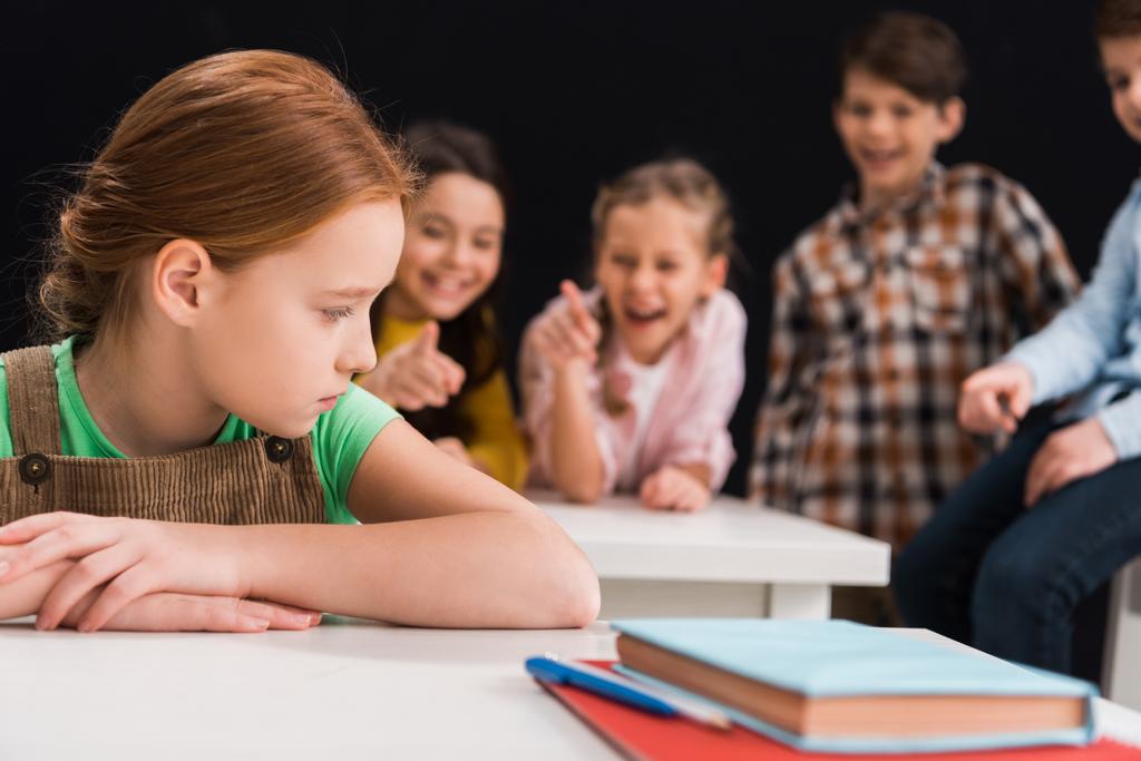 selective focus of upset schoolkid sitting near classmates laughing and pointing with fingers isolated on black, bullying concept  - Photo, Image