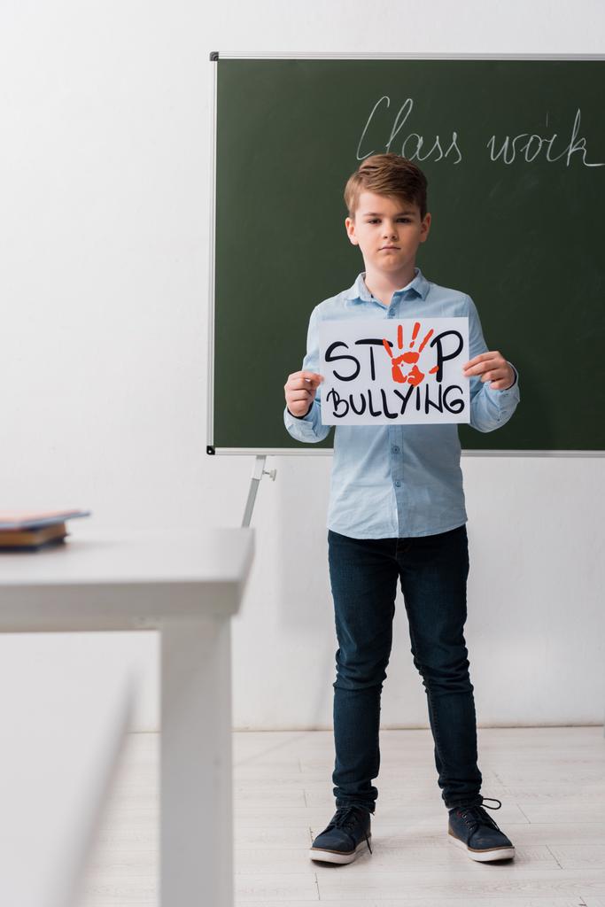 selective focus of schoolchild holding placard with stop bullying lettering in classroom  - Photo, Image