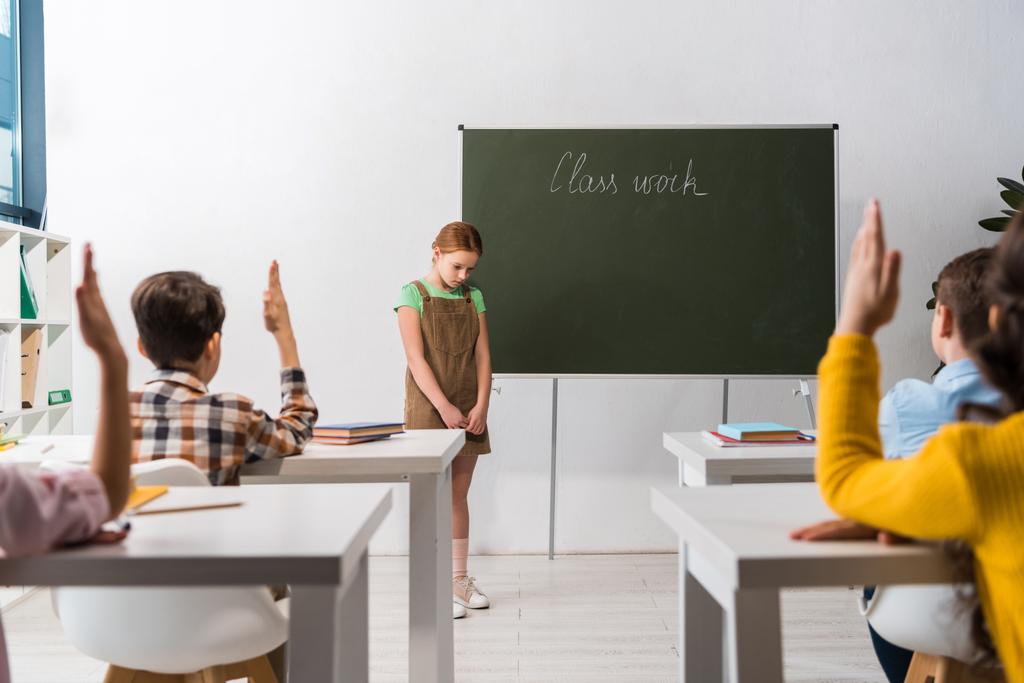 foco seletivo de estudante em pé perto de quadro-negro com letras de trabalho de classe e colegas de classe com as mãos levantadas
  - Foto, Imagem