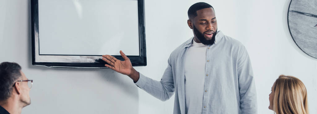 plano panorámico de sonriente hombre de negocios afroamericano apuntando con la mano a la televisión y hablando con colegas
  - Foto, imagen