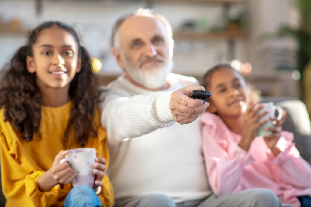 Twee donkergekleurde meisjes kijken tv met hun opa en glimlachen - Foto, afbeelding