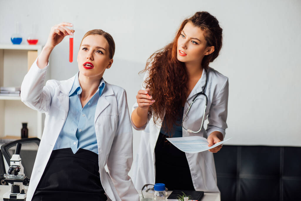 piękne i seksowne pielęgniarki patrząc na probówkę z czerwonym płynem - Zdjęcie, obraz