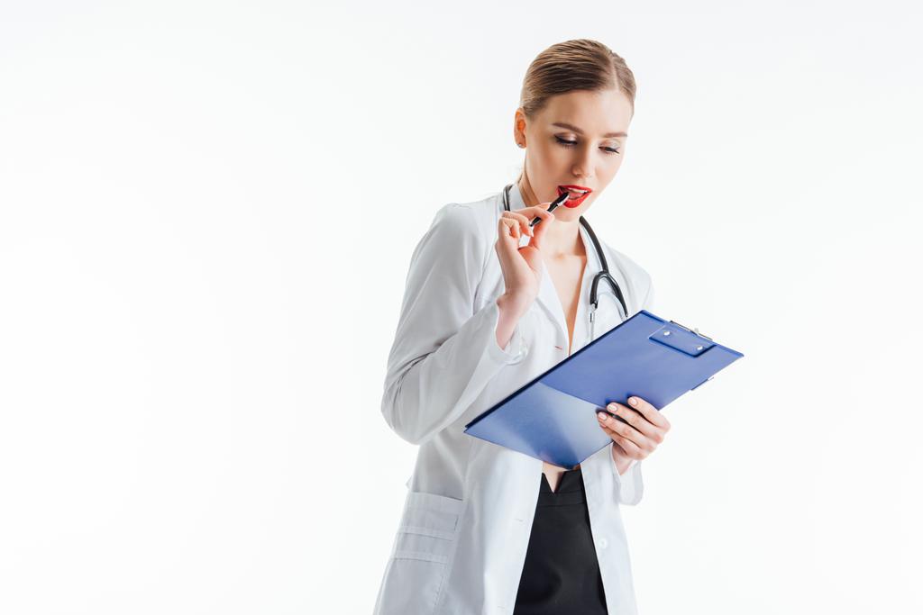 zamyšlené a sexy zdravotní sestra v bílém plášti při pohledu na předpis při držení schránky a pero v blízkosti červené rty izolované na bílém  - Fotografie, Obrázek