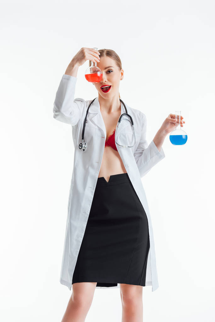 χαρούμενη και σέξι νοσοκόμα σε λευκό παλτό κρατώντας φιάλες με κόκκινο και μπλε υγρό απομονώνονται σε λευκό  - Φωτογραφία, εικόνα