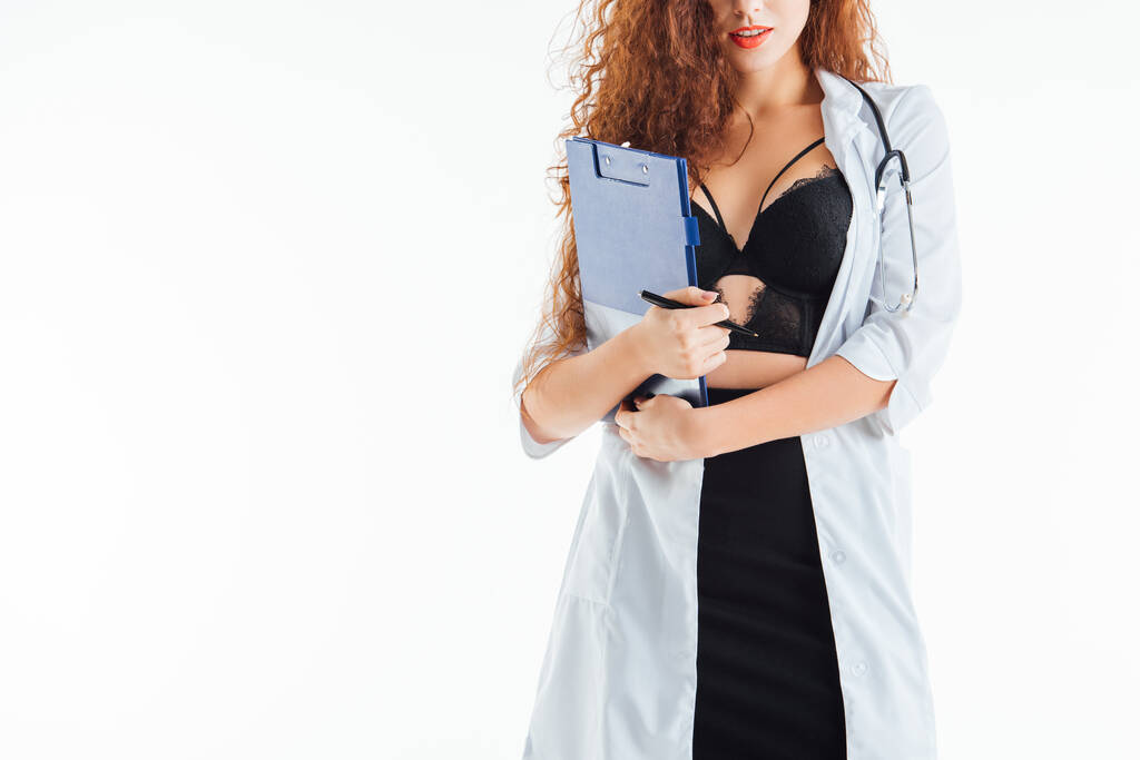καλλιεργημένη άποψη του σγουρά και σέξι νοσοκόμα κρατώντας πρόχειρο απομονωμένο σε λευκό  - Φωτογραφία, εικόνα